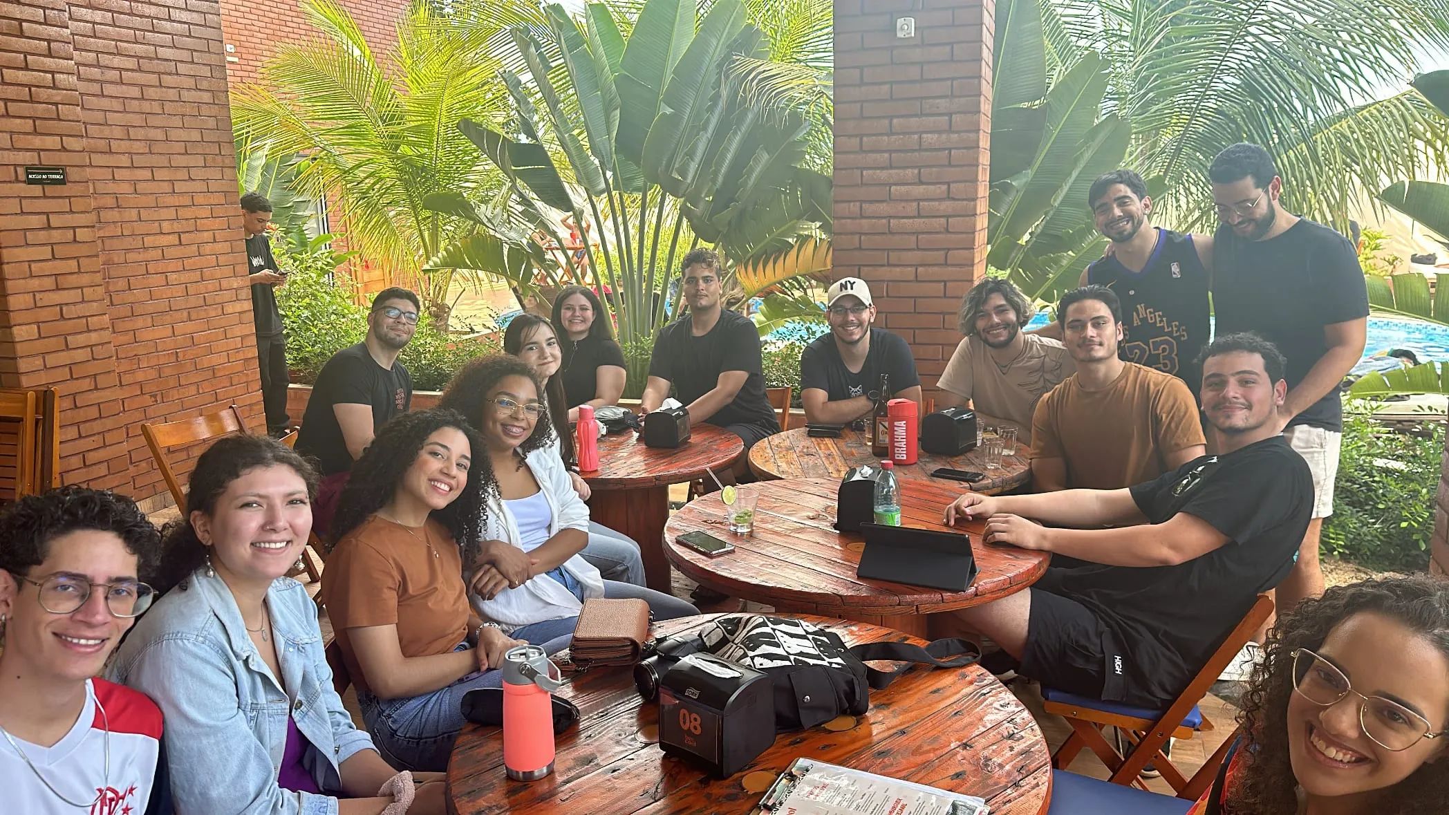 Participantes sorrindo ao redor de mesas de madeira ao ar livre, no 1º encontro presencial